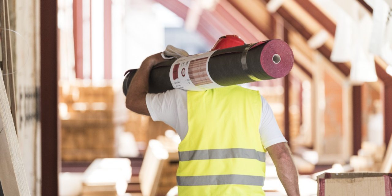 Urban roofer carrying underlay sheet on his shoulder in renovated attic wearing hard hat and safety jacket