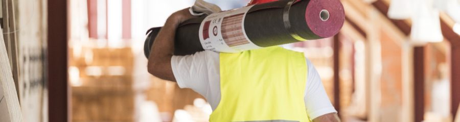 Urban roofer carrying underlay sheet on his shoulder in renovated attic wearing hard hat and safety jacket
